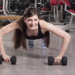 Sportlerin im Fitnesscenter rollt sich mit Kurzhanteln auf dem Boden und trainiert so Ihre Bauchmuskeln.
