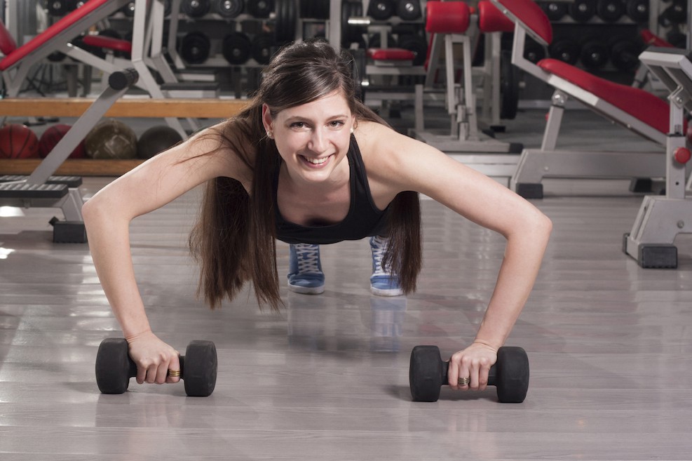 Sportlerin im Fitnesscenter rollt sich mit Kurzhanteln auf dem Boden und trainiert so Ihre Bauchmuskeln.